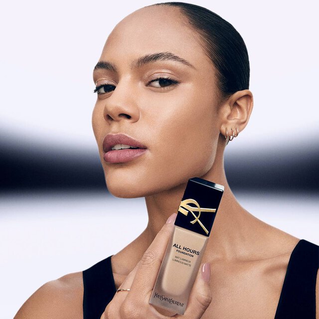 A person with sleek, pulled-back hair poses confidently, holding a bottle of Yves Saint Laurent All Hours Foundation. They have a natural makeup look, showcasing smooth skin and subtle lip color, set against a minimalist background.