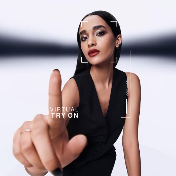 Vrouw wijst naar voren, interactief met virtuele pas-scherm. Grafische overlay toont "VIRTUAL TRY ON" en "LOOK XXXX" rond haar gezicht. Ze draagt een zwart vest​