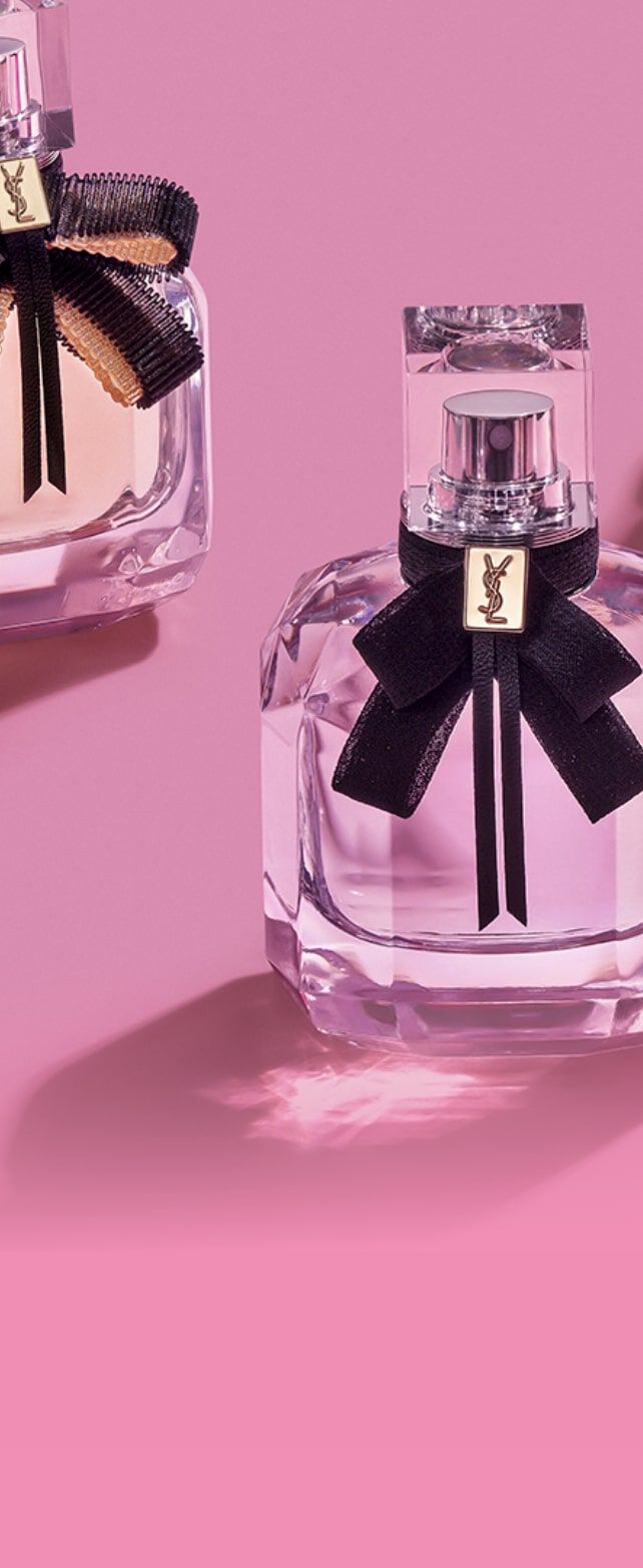 Drie flacons parfum op een roze achtergrond, elk met een zwarte strik en een verschillende kleur vloeistof: lichtroze, paars en diep paars.