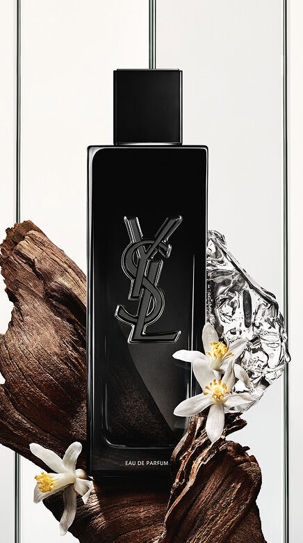 Een zwarte Yves Saint Laurent Eau de Parfum fles met het YSL-logo, omringd door donkere houtstukken, delicate witte bloemen en een verkreukeld zilveren, reflecterend object.