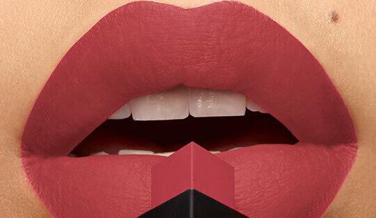 Close-up of full lips wearing a matte pink lipstick, with a geometric lipstick case positioned in front of the mouth.