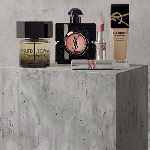 Promotionele display van YSL Beaute make-up producten, waaronder mascara, een gewatteerd oogschaduwpalet en lippenstiften tegen een betonnen achtergrond.