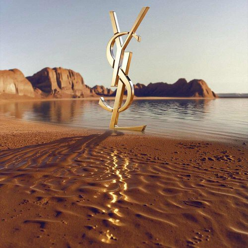 Een groot, iconisch 3D gouden YSL (Yves Saint Laurent) logo op een woestijnachtige kustlijn. Het gepolijste gouden monogram reflecteert het zonlicht en staat voor een kalm wateroppervlak met spectaculaire rode rootsformaties en woestijnbergen op de achtergrond onder een heldere hemel.