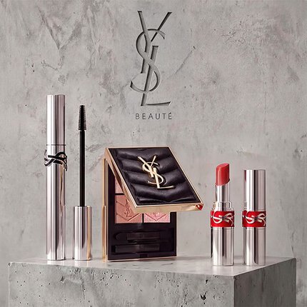Promotionele display van YSL Beaute make-up producten, waaronder mascara, een gewatteerd oogschaduwpalet en lippenstiften tegen een betonnen achtergrond