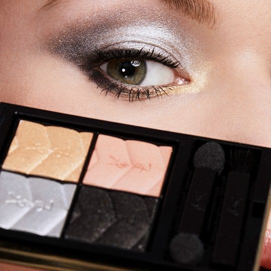 Close-up of a person's eye with expertly applied silver and gray eyeshadow, highlighting a makeup palette featuring shades of beige, silver, and black.