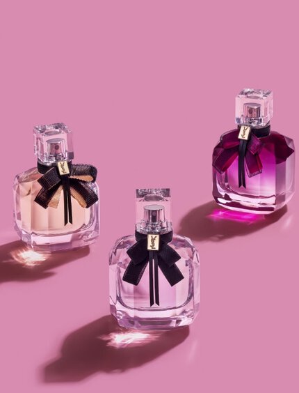 Three elegant perfume bottles with black ribbons, displayed on a pink background. The bottles are in varying shades, including light pink and purple, casting soft shadows.