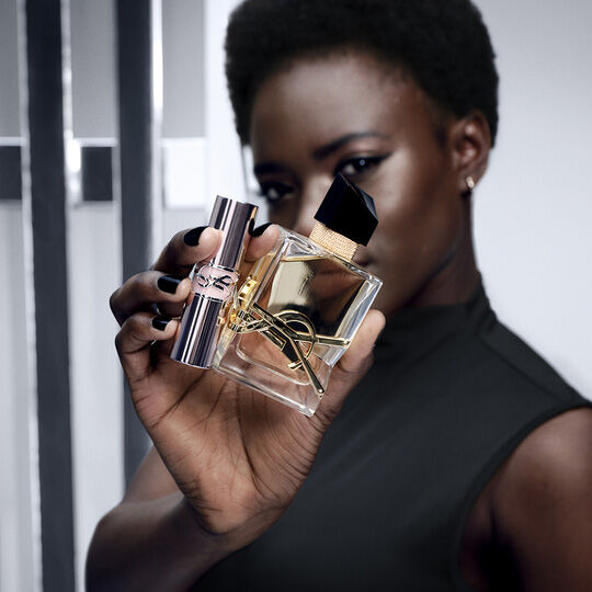 Een vrouw met een donkere huid en kort haar houdt een flesje Yves Saint Laurent parfum en een bijpassende lipstick vast, beide met het iconische YSL-logo.