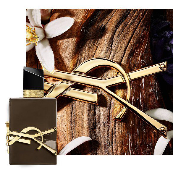Libre Santal Couture geglazuurde bruine flacon op sandelhoutschors met gouden Cassandre en witte bloemen.