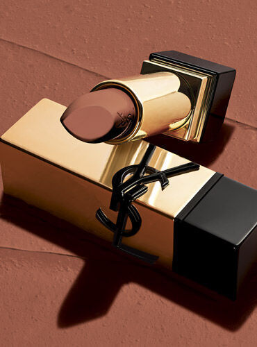 A luxurious gold lipstick bullet with a sleek black base, resting on a textured brown surface. The lipstick is a rich, deep shade.