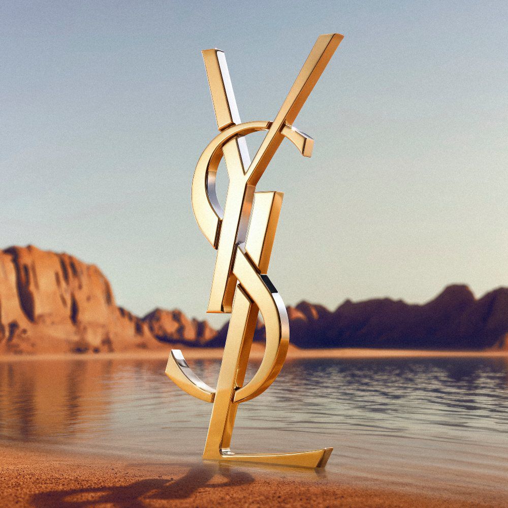 Een groot, iconisch 3D gouden YSL (Yves Saint Laurent) logo op een woestijnachtige kustlijn. Het gepolijste gouden monogram reflecteert het zonlicht en staat voor een kalm wateroppervlak met spectaculaire rode rootsformaties en woestijnbergen op de achtergrond onder een heldere hemel.