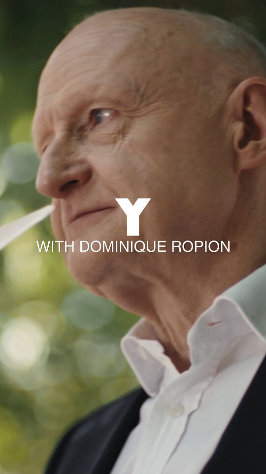 Een kale man in een zwart pak en wit overhemd houdt verschillende witte papieren stroken, die gewoonlijk worden gebruikt voor geurtesten, tegen zijn neus alsof hij een geur evalueert. Hij kijkt peinzend, met een groene, wazige achtergrond van gebladerte en bomen. Over de afbeelding staat de grote witte tekst "Y" met daaronder de kleinere tekst "WITH DOMINIQUE ROPION".