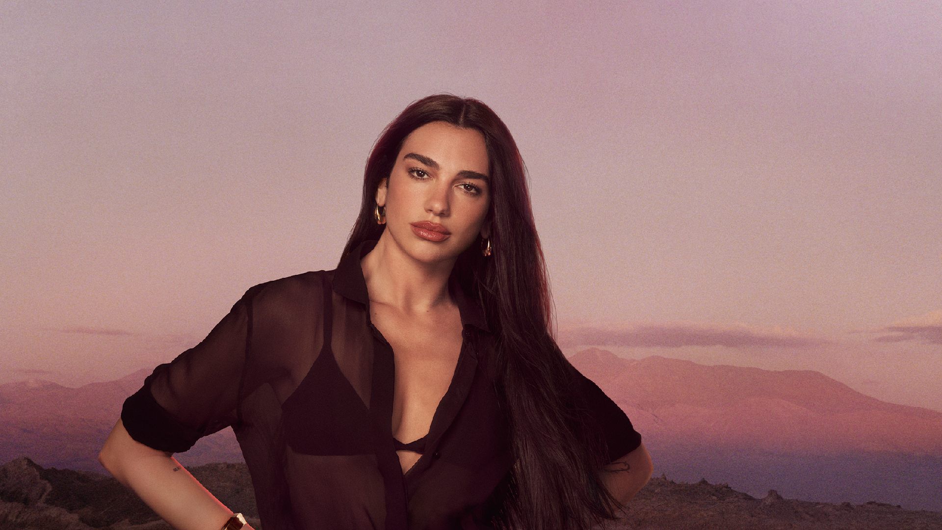 Dua Lipa poseert in een zwart transparant overhemd tegen een berglandschap bij zonsondergang.