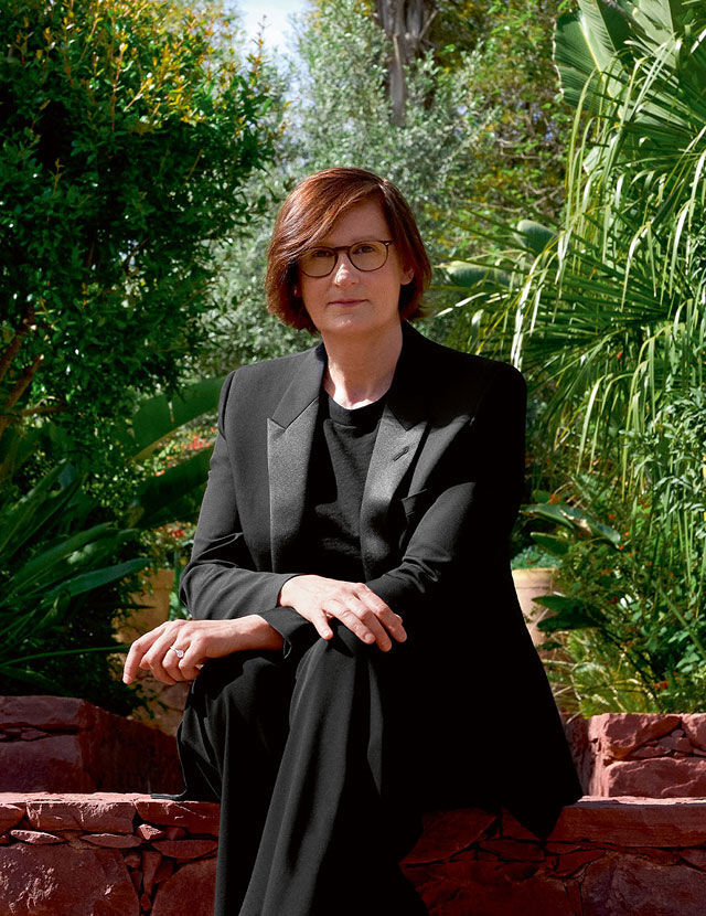 Een vrouw met roodbruin haar en een bril zit op een stenen muur en draagt een zwart pak. Ze kijkt de kijker aan, met een weelderige groene tuin op de achtergrond.