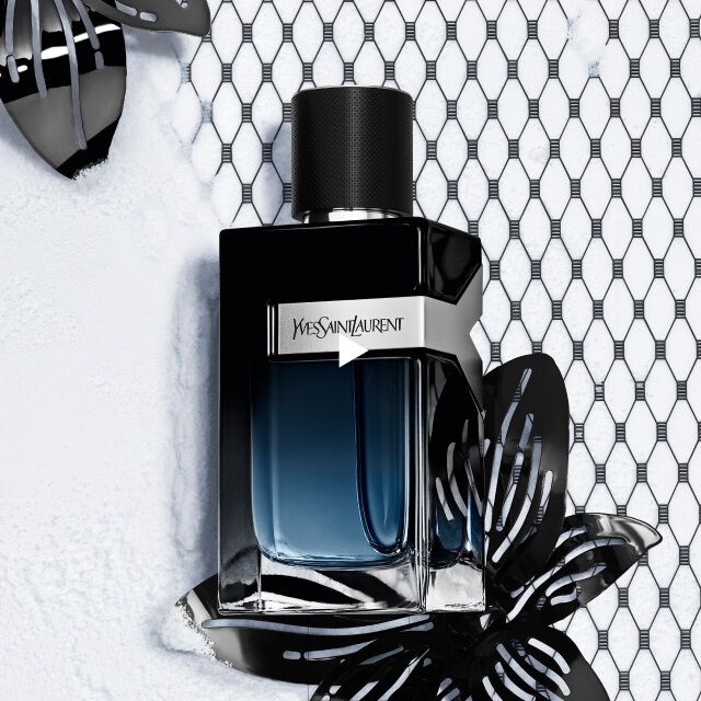 A dark, sleek perfume bottle with a gradient blue and black design is surrounded by black decorative butterflies and a textured net background. The label reads "Yves Saint Laurent."