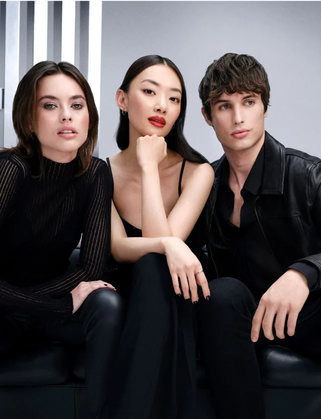 Drie stijlvolle jonge modellen in zwarte kleding zittend op een zwart leren bank tegen een grijze studio-achtergrond.