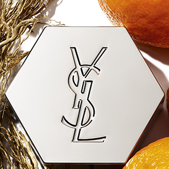 A silver hexagonal compact with the YSL logo, surrounded by vibrant orange slices and intertwined roots.