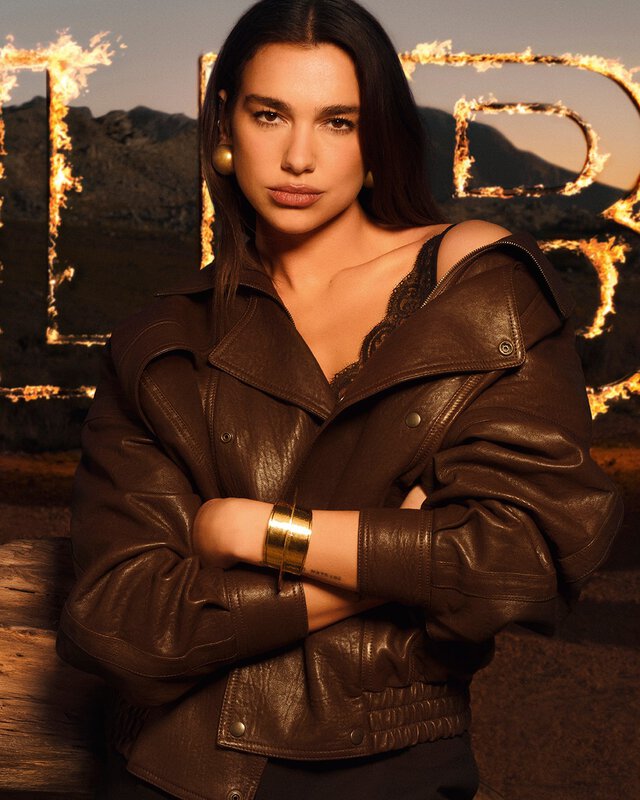 Dua Lipa in een bruine leren jas met grote vlammende LIBRE-letters in een rauw woestijnlandschap.
