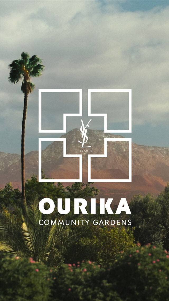 Een landschap met palmbomen, groen gebladerte en bergen onder een bewolkte hemel. Een wit YSL Beaute-logo en de tekst "OURIKA COMMUNITY GARDENS" zijn eroverheen gelegd.