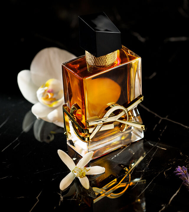 A stylish perfume bottle with a black cap and gold accents sits on a dark marble surface, surrounded by white flowers.
