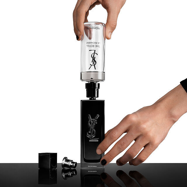 De handen van een persoon vullen een zwarte Yves Saint Laurent (YSL) parfumfles bij met een transparante navulcartridge waarop "THE REFILL LA RECHARGE" staat.