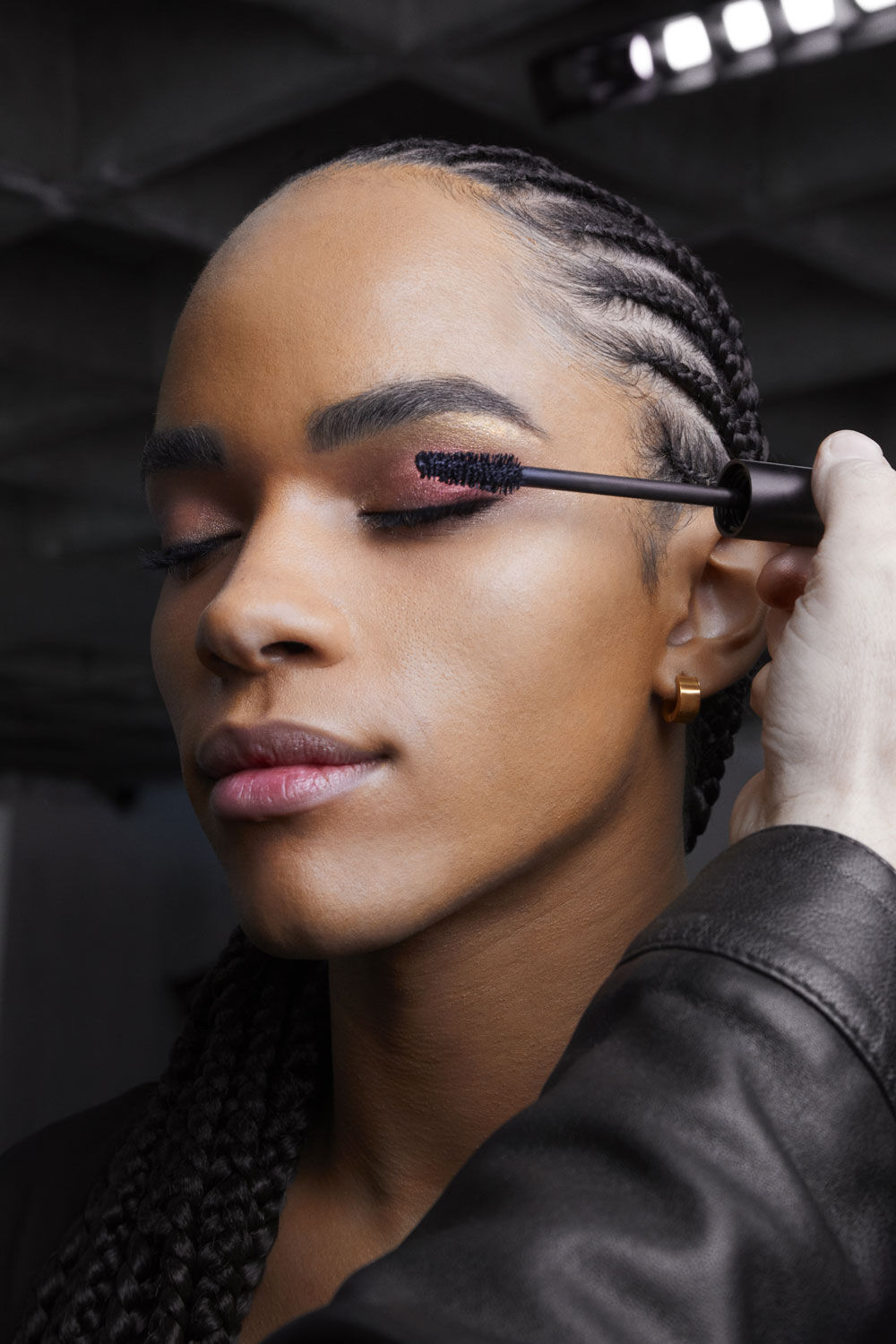 A close-up of a person with braided hair applying mascara to their eyelashes, with a soft focus on their face. The setting appears to be a studio with neutral lighting.