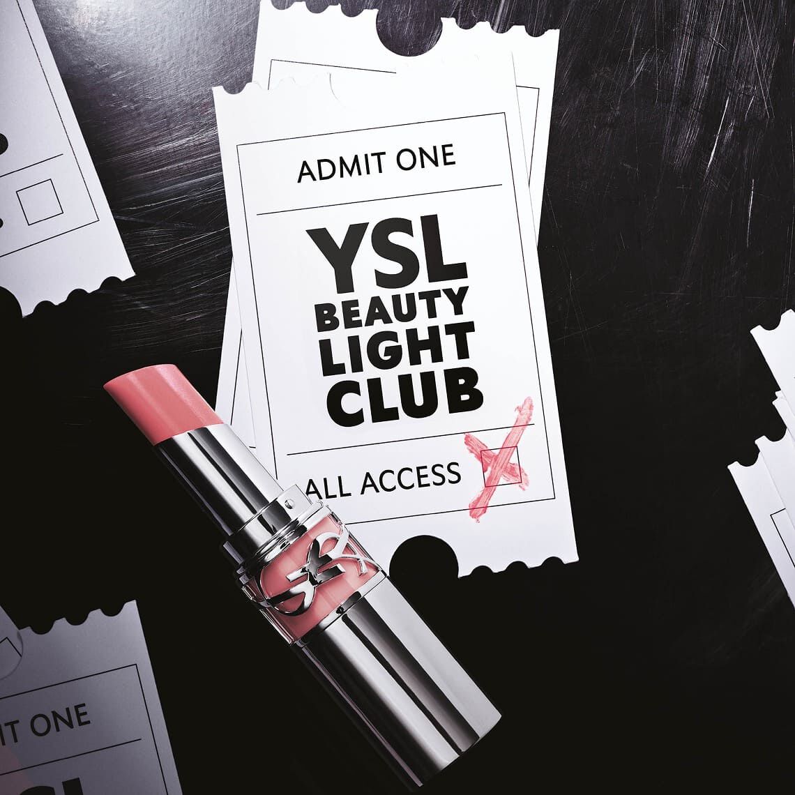 Een lichtroze lippenstift met een glanzende textuur ligt op twee licht overlappende witte evenementtickets. Het bovenste ticket vermeldt bovenaan "ADMIT ONE" en in het midden, in grote zwarte letters, "YSL BEAUTY LIGHT CLUB", met daaronder "ALL ACCESS". Een rood kruis is in de rechteronderhoek van het ticket gekrabbeld, alsof het gevalideerd is. De achtergrond is donker, wat een getextureerd oppervlak suggereert, en aan de zijkanten zijn gescheurde ticketranden zichtbaar, wat duidt op de aanwezigheid van meerdere andere verspreide tickets. Het geheel geeft de indruk van een exclusieve uitnodiging voor een glamoureus evenement.
