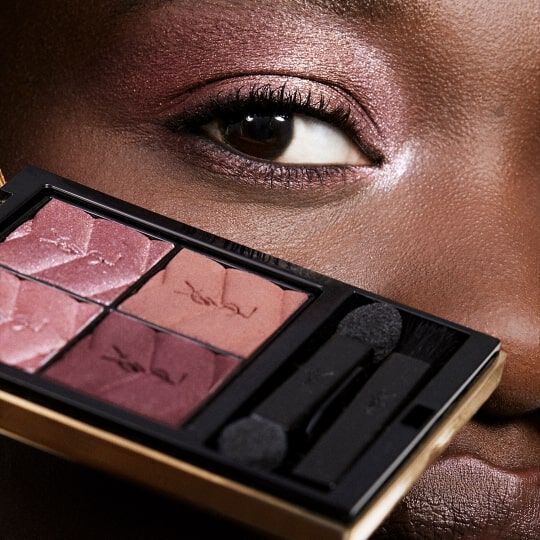 A close-up of a person's eye, featuring a makeup palette with shades of pink and brown positioned in front of their face. The eye showcases eyeshadow that complements the palette colors.