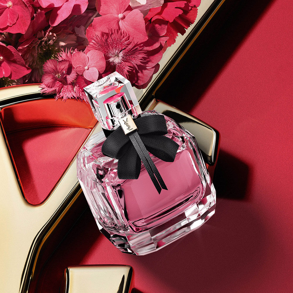 YSL Mon Paris Eau de Parfum fles met roze geur en een zwarte strik, gestyled met roze bloemen op een rode achtergrond met een gouden YSL-logodetail.