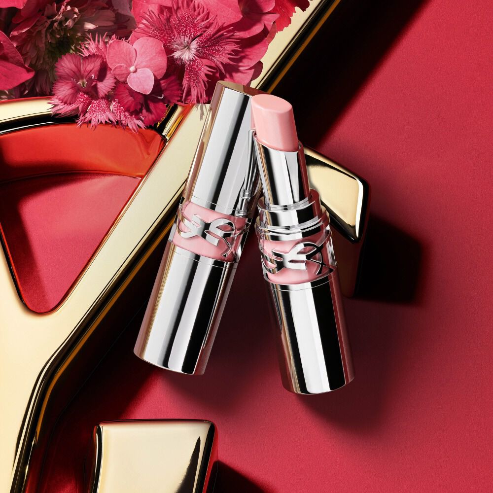 YSL Loveshine lippenstift kleur 44 in zilveren verpakking, open getoond met een zachte roze kogel naast de gesloten buis, gestyled met roze bloemen op een rode achtergrond met gouden YSL-logodetails.