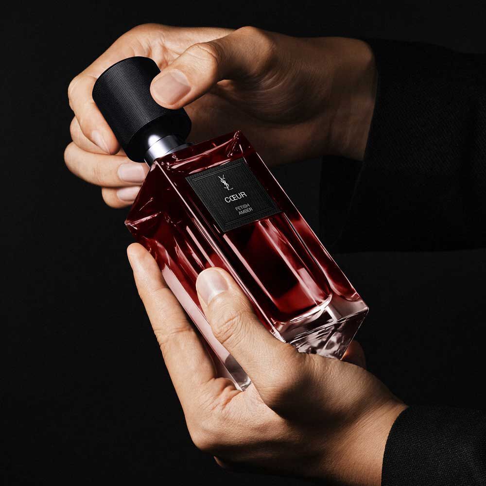 Handen houden de COEUR Eau de Parfum robijnrode fles vast om de YSL Art of Application en couture-ontwerp te demonstreren.