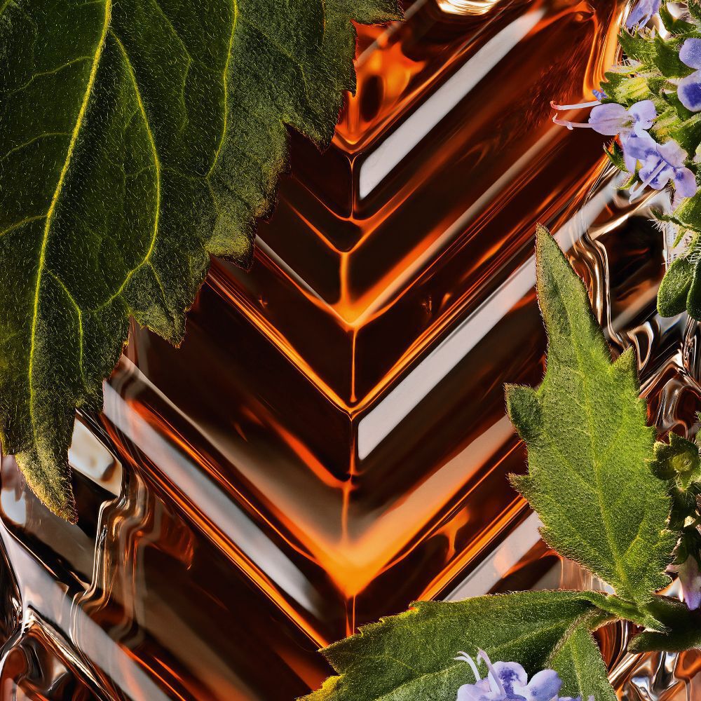 Abstracte close-up van YSL Tuxedo Extrait de Parfum het getextureerde glas van de fles, omlijst door levendige patchoulibladeren en bloemen.