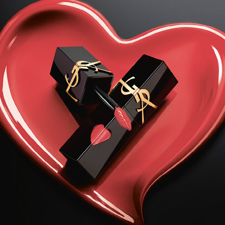 Twee matte zwarte YSL lipstainflacons met roze hartvormige uitsparingen en gouden logo’s, tentoongesteld in een bijpassend gebeeldhouwd mat roze hart op een zwarte achtergrond.