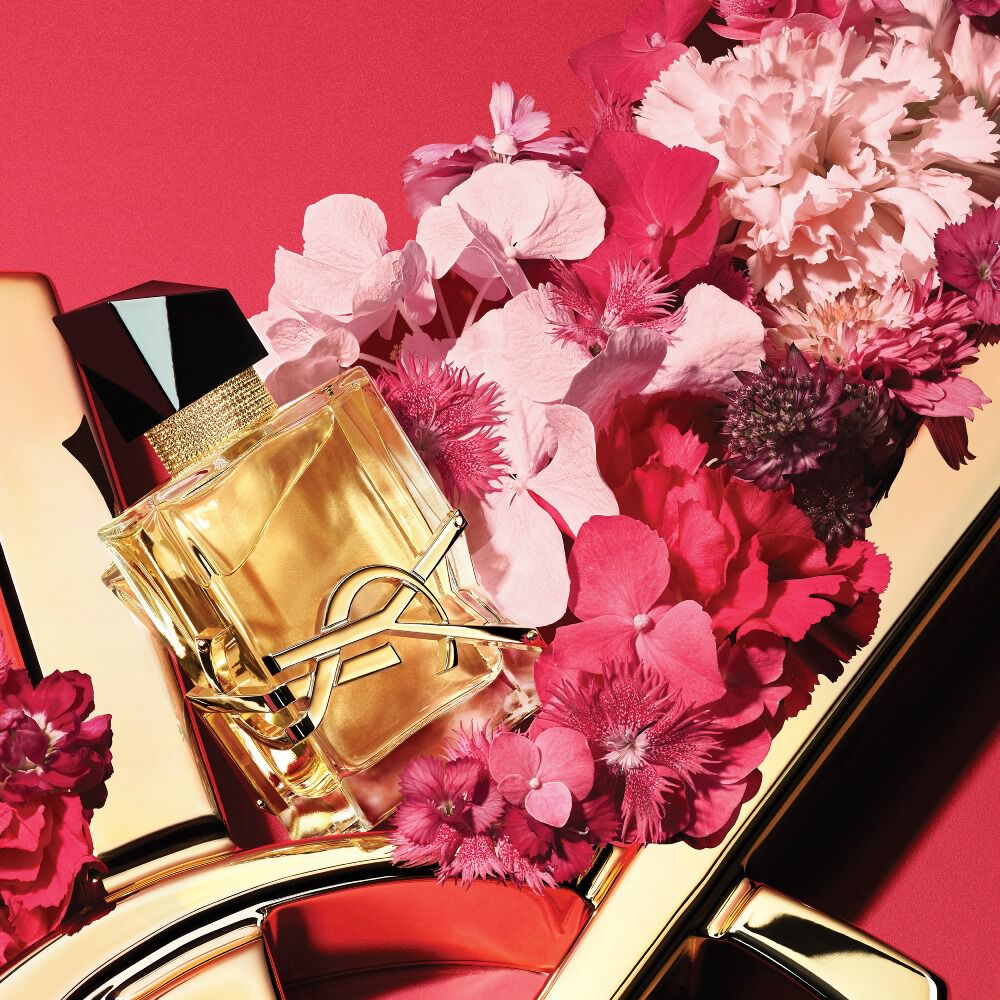 YSL Libre Eau de Parfum fles met gouden YSL-logo, gestyled met roze en rode bloemen op een rode achtergrond met een gouden YSL-logodetail.
