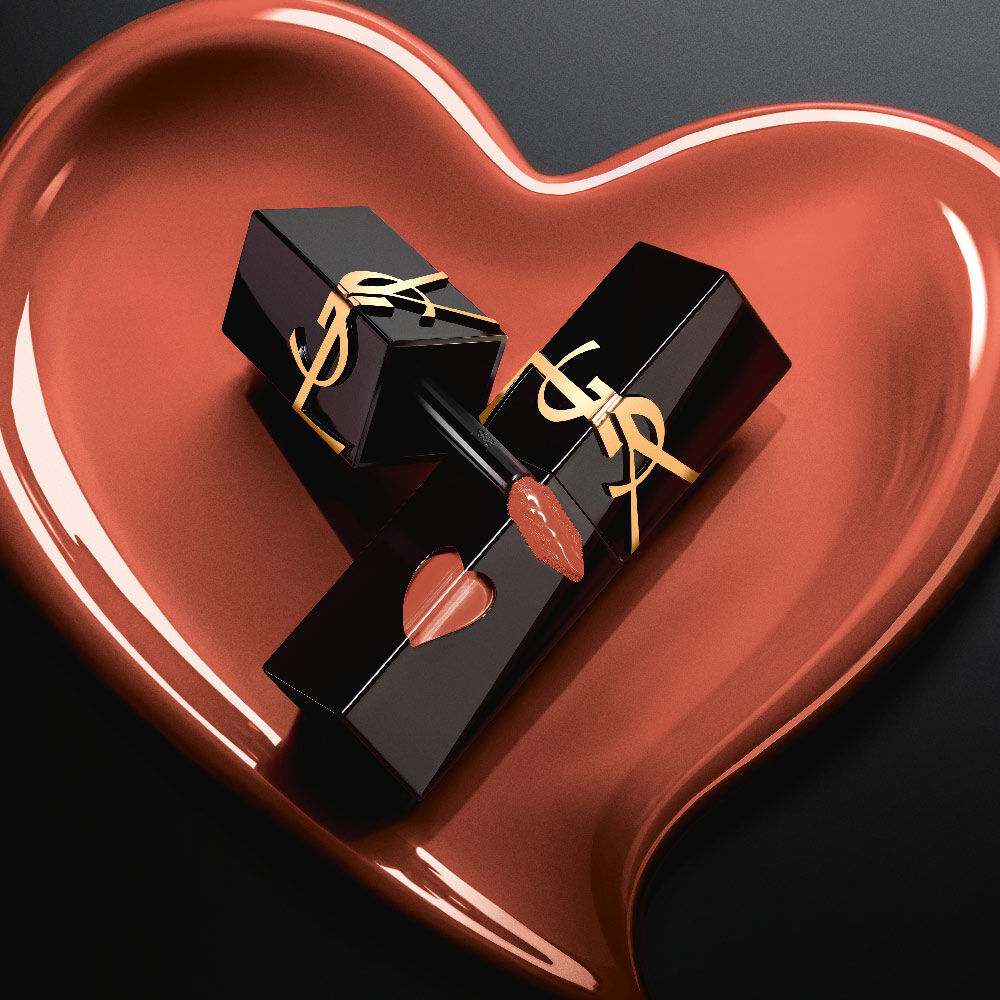 Twee matte zwarte YSL lipstainflacons met roze hartvormige uitsparingen en gouden logo’s, tentoongesteld in een bijpassend gebeeldhouwd mat roze hart op een zwarte achtergrond.