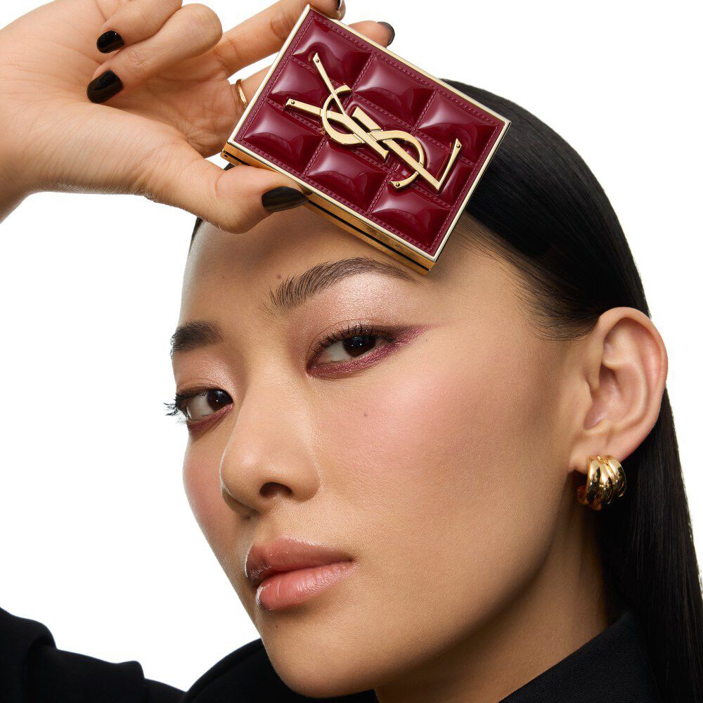 Model in een zwart pak en een glamoureuze look, gezeten in een feestelijke kaarsverlichte setting met YSL cadeauverpakkingen.