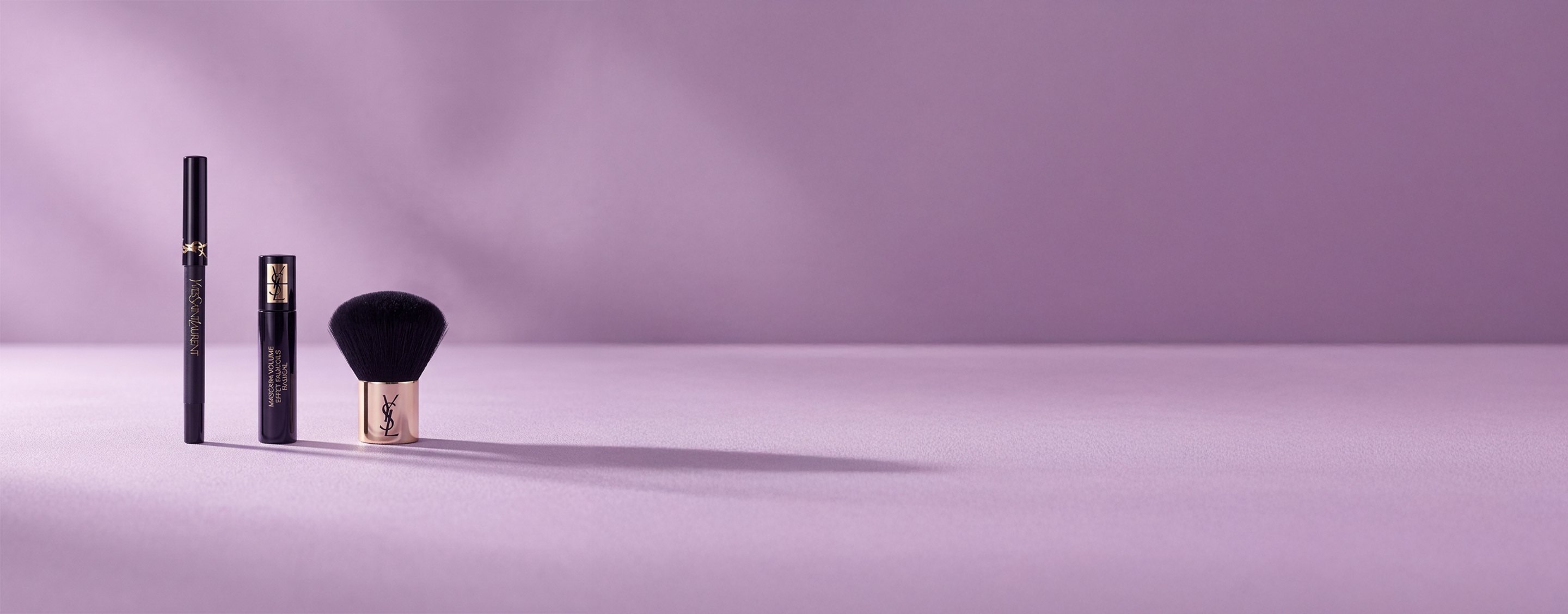 Drie Yves Saint Laurent-schoonheidsproducten, waaronder een mascara en een kabuki-kwast, op een zachte lavendelkleurige achtergrond.