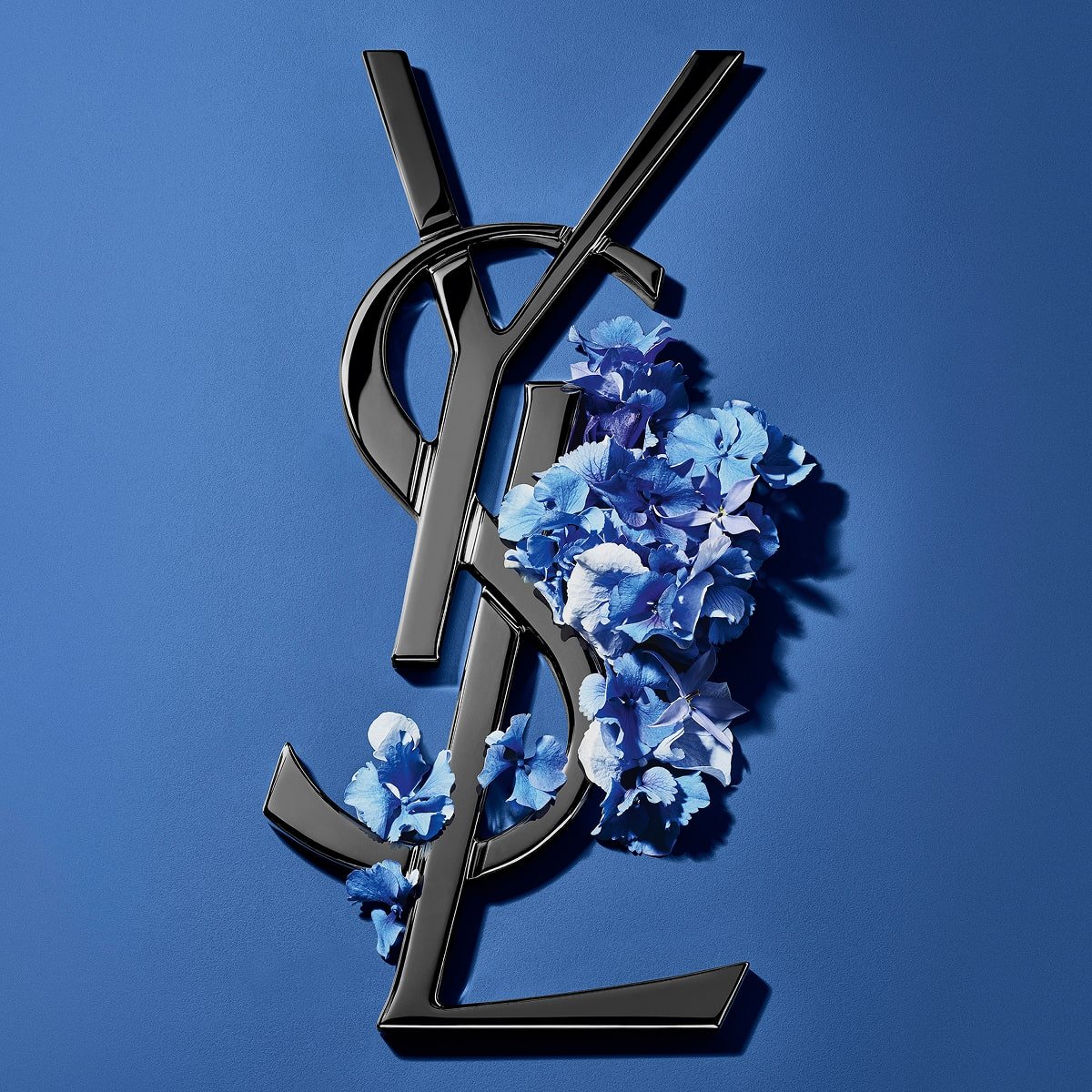 Zwart YSL Cassandre-logo versierd met levendige blauwe voorjaarsbloemen, tegen een serene blauwe achtergrond.