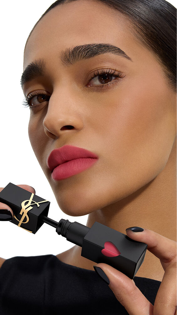 Model met een middelmatige huidskleur en een levendige roos-matte lippenkleur houdt de zwarte fles van YSL THE INKS BLUR lippenvlek met het roze hart vast.