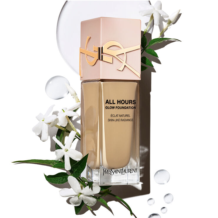 Fles YSL All Hours Glow Foundation met 'Éclat Naturel Skin-Like Radiance' omringd door witte jasmijnbloemen en heldere waterdruppels, wat natuurlijke ingrediënten en hydratatie suggereert.