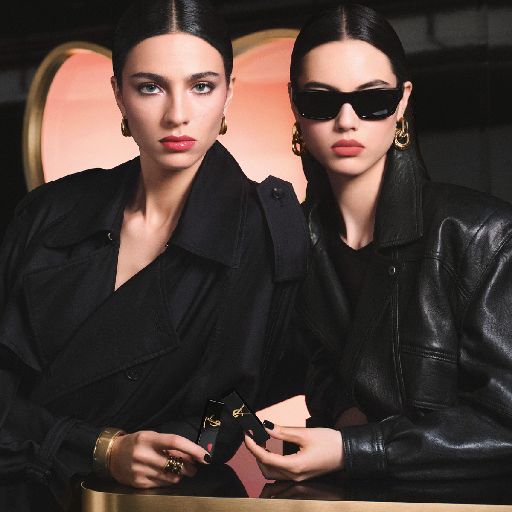 Twee stijlvolle modellen in zwarte trenchcoat en leren jasje, met YSL-lipsticks voor een grote roze hartvormige achtergrond, tonen een glanzende rode liplook en een zacht-matte roze liplook.