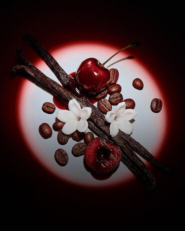 Close-up van sappige rode kersen, jasmijnbloemen, vanillepeulen en koffiebonen, uitgelicht door een dramatische spot voor Black Opium Eau de Parfum Over Red.