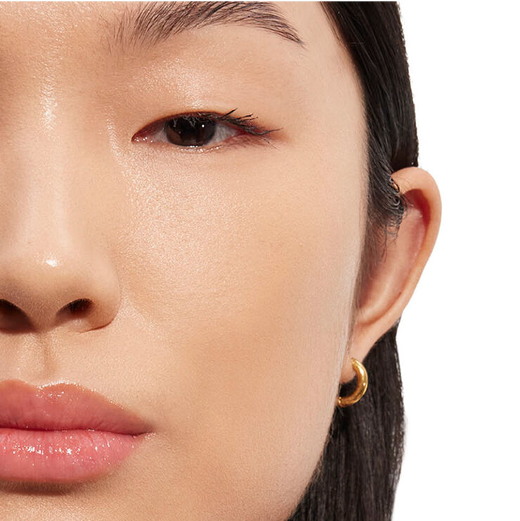 Gedetailleerde close-up van het gezicht van een persoon, gericht op het oog- en wanggebied met natuurlijk ogende make-up en een gouden creool, die de helderheid van de huid demonstreert.