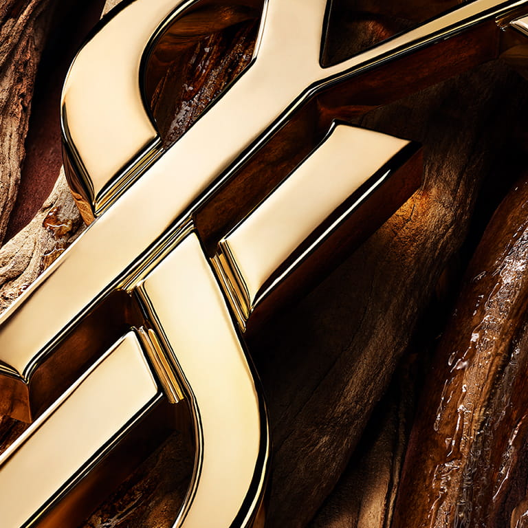 Close-up of the YSL Cassandre logo in polished gold set against warm vanilla pods from Madagascar.