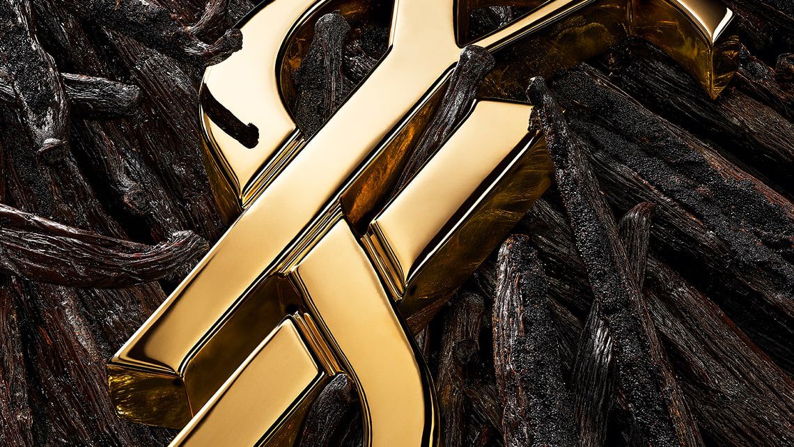 Nahaufnahme des goldenen YSL-Logos, platziert zwischen Vanilleschoten, weißen Blüten und funkelnden goldenen Akzenten.