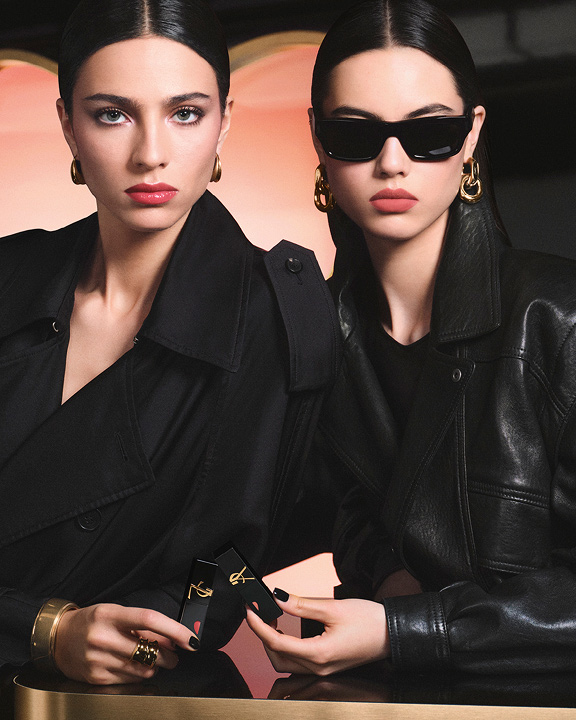 Twee stijlvolle modellen in zwarte trenchcoat en leren jasje, met YSL-lipsticks voor een grote roze hartvormige achtergrond, tonen een glanzende rode liplook en een zacht-matte roze liplook.