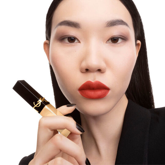 Hand houdt een zwart‑gouden YSL lippenstift met vierkante punt bij de kin.