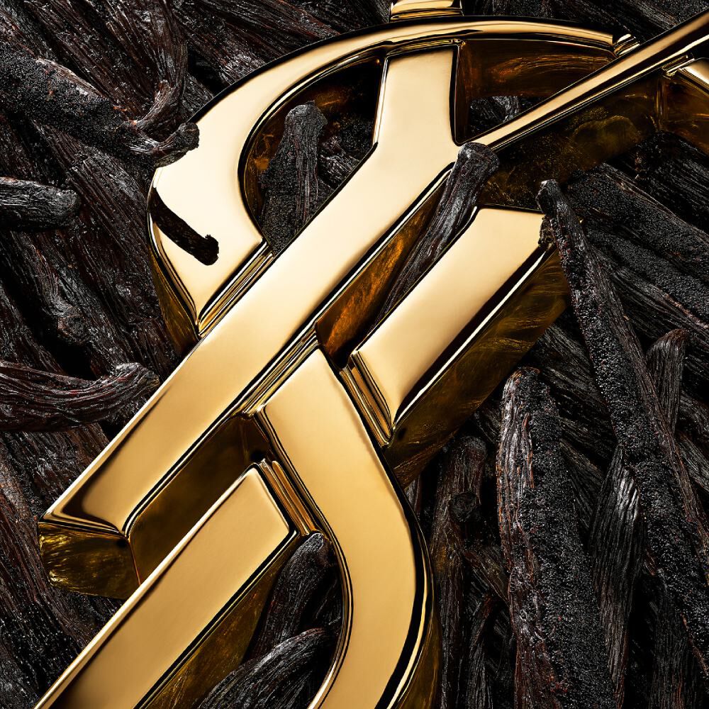 Zeer gestileerde close-up van het gouden YSL-logo over donkere vanillepeulen, waarbij de texturen worden geaccentueerd.