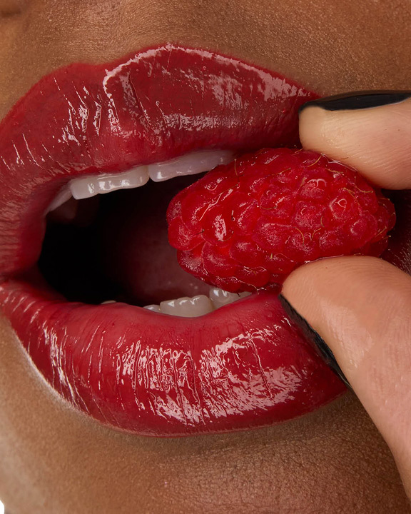 Close-up van de lippen van model Zahara, verfraaid met YSL LOVESHINE Lipstick in de kleur Berry Crush, tint 219, terwijl ze een verse framboos vasthoudt.