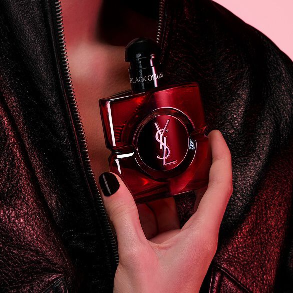 Close-up van een hand met zwarte nagels die de flacon Black Opium Eau de Parfum Over Red vasthoudt tegen een zwarte leren jas.
