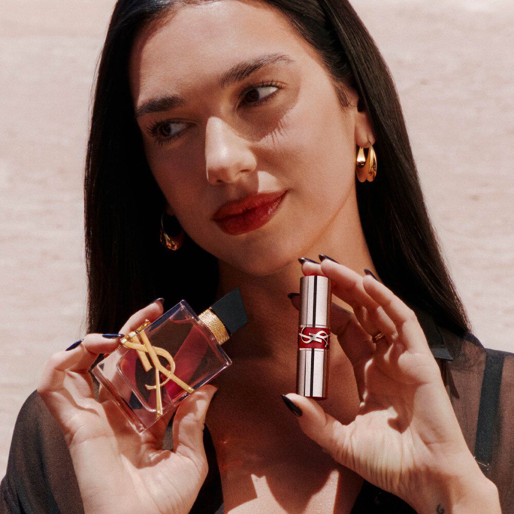 Dua Lipa houdt Libre Eau de Parfum Berry Crush en een YSL Loveshine vast, met rode lippen en gouden oorbellen.